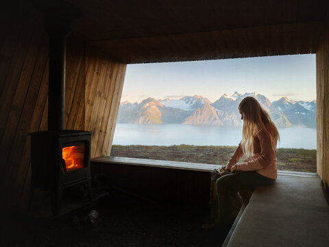 Traveler woman warming up beside fireplace in mountain cabin with panoramic window view fjord and Lyngen Alps landscape in Norway, cozy shelter hiking adventure vacations solo slow travel lifestyle
