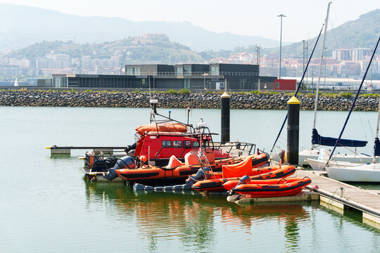 Red rescue boats and inflatable lifeboats docked at a harbuor pier alongside sailboats with coastal buildings in the background.