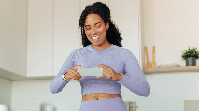 Smiling Black Woman Engaging With Smartphone While Standing In Modern Kitchen, Enjoying Interaction With Digital Content, Happy Emotion Visible On Face. Weight Loss Concept.