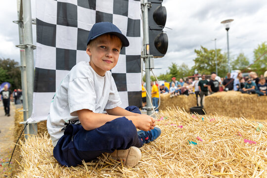 Boy watches soap box racing from hay bales beside checkered flag, enjoying outdoor community event in Germany. Young spectator rests near finish area while crowd gathers around downhill race course