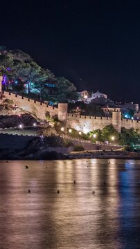 Panoramic view of the medieval walled town of Tossa de Mar in Spain at night, with the ancient castle and its colorful lights beautifully reflected on the calm Mediterranean Sea