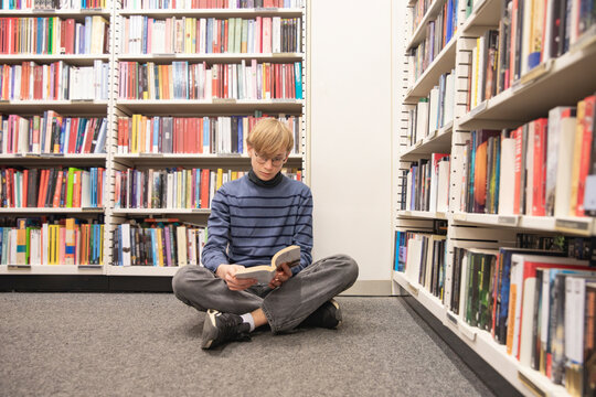 young student sitting cross legged on library floor reading book