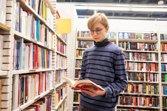 A young boy wearing glasses standing and reading interesting book near bookshelves