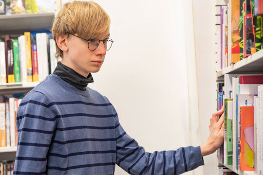 young male student wearing glasses choosing book from bookshelf in bookstore or library