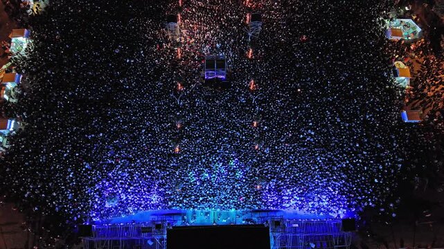 Aerial top view of crowd at night music concert. People gathered at festival outdoor stage with lights and screens, 4k