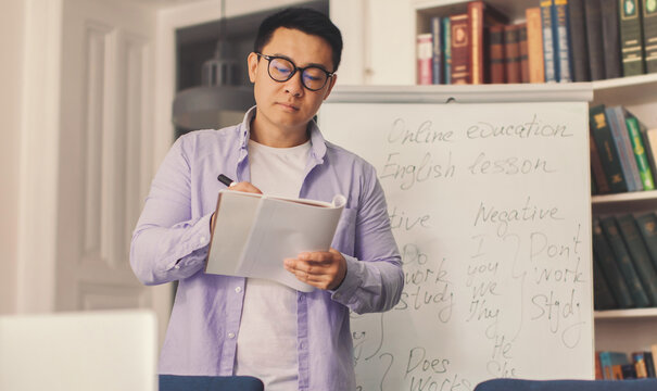 Focused Asian Male Teacher Taking Notes While Conducting An Online English Lesson At Home, Standing In Front Of A Whiteboard Filled With Writing, Emphasizing Learning And Education Concept.