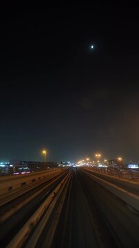 Front POV shows a train moving forward along an elevated metro railway at night under the moon, passing illuminated streetlights and city traffic