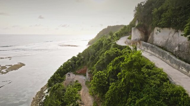 Drone aerial view flight over scenic road carved into the steep cliffside, overlooking the vast Indian ocean and Green Bowl Beach at low tide, surrounded by lush green vegetation in Bali, Indonesia