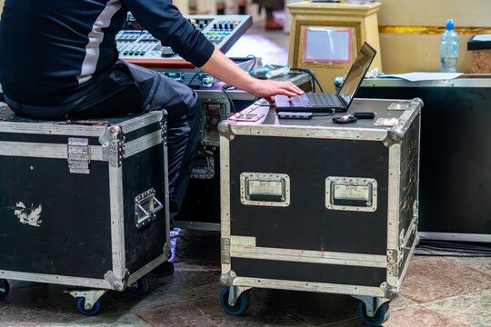 Man sitting on black transport flight case operating laptop placed on adjacent equipment road case near sound mixing console, indoor photography shot under artificial lighting.