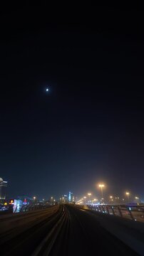 Camera travels along an elevated metro track at night under a crescent moon, moving toward distant city lights and illuminated streetlamps