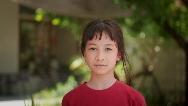 Portrait of adorable Asian kid looking directly at the camera with a gentle smiling face in a red shirt standing outdoors with beautiful green bokeh background. Childhood happiness lifestyle concept.