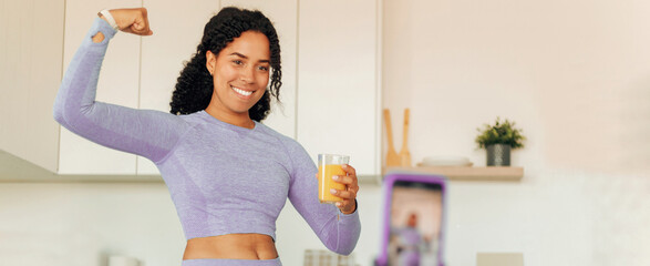 Smiling Black Woman With Positive Emotion Flexing Muscles While Holding A Glass Of Juice At Home In...
