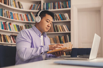 Fototapeta premium Focused Asian Male Student Concentrating On Reading Book While Using Laptop At Home Office. Engaging In Study Or Work Session In A Modern Workspace, Wearing Headphones, Productive Environment.