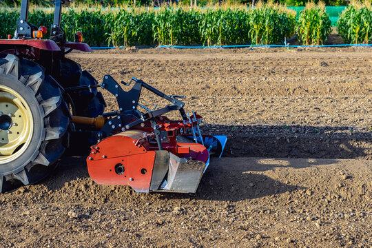 Agricultural tractor standing with front blade and rear rotary tiller on tilled agricultural field during land preparation for farming with trees and cornfield in background. Agricultural machinery.