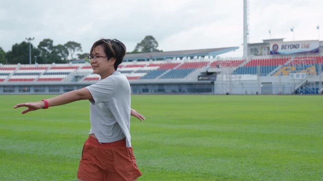 Authentic shot of an active Asian woman stretching her arms on a green soccer field. This empty stadium with colorful seats provides excellent copy space for your sports backgrounds.
