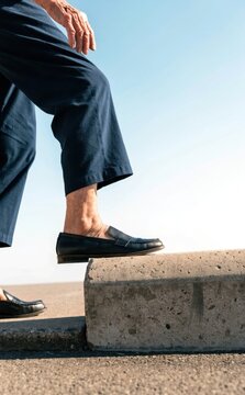 Senior functional fitness concept. Person stepping up on a concrete block wearing black pants and black loafers under a clear blue sky.