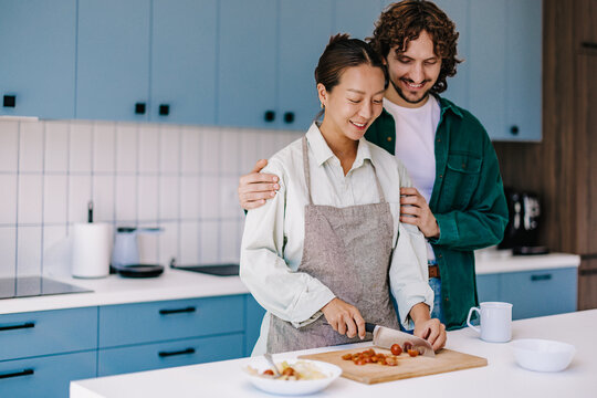 Happy Couple Cooking Together In Modern Home Kitchen, Woman Chopping Vegetables While Partner Embraces Smiling