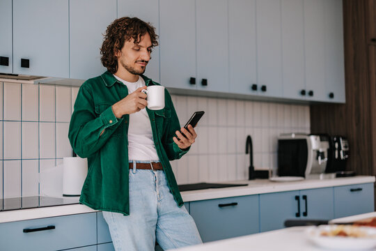 Casual Man Drinking Coffee While Checking Smartphone in Modern Kitchen &mdash; Remote Work Lifestyle