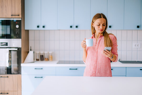 Young Woman In Modern Kitchen Holding Coffee Mug And Checking Smartphone, Casual Morning Routine