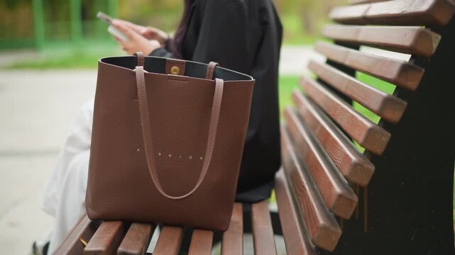 Close up of brown leather tote bag with FASHION embossed on it placed centrally in focus. person sits behind bench using smartphone while wearing dark clothing and white pants only their