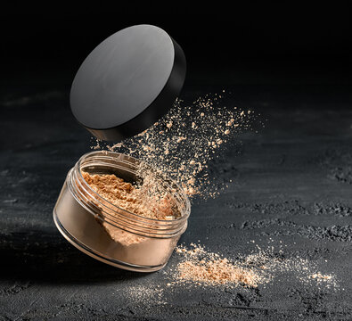 Close-up open loose face powder jar with puff releasing fine cosmetic powder cloud over dark textured tabletop