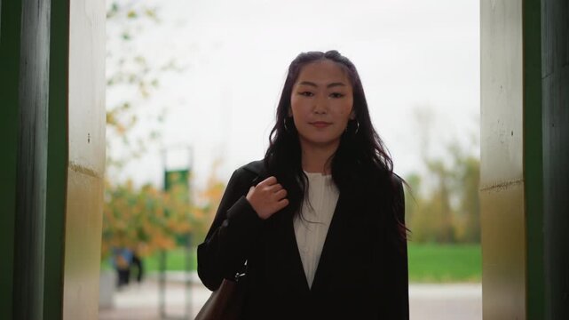Close up of young woman walking outdoors in urban park setting holding her blazer lapel with one hand and carrying brown bag. Woman wears black coat over white blouse against backdrop