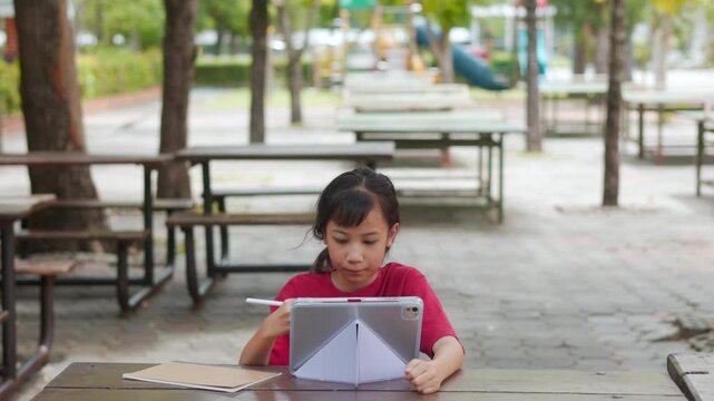 Asian kid doing homework on digital tablet at outdoor campus table. The selective focus shows the student enjoying active e-learning, highlighting modern education technology and tech-savvy childhood.