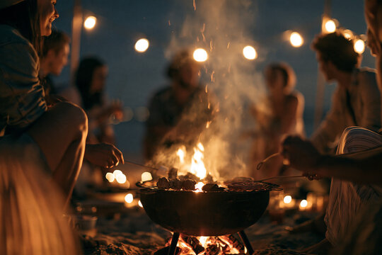 Group of friends enjoying outdoor barbecue at night, sitting around fire pit on sandy beach, warm lights in background, relaxed atmosphere, summer evening gathering, joyful and cozy mood