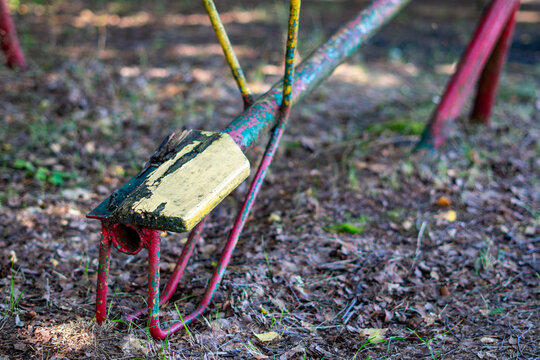 PRL-era playground featuring a close-up of an old, worn seesaw with a damaged wooden seat and peeling paint, red, green, and yellow metal tubes, forest ground with pine needles and dry leaves.