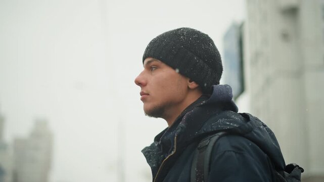 Close up of man wearing winter clothing and carrying backpack standing outdoors in urban environment with tall buildings. scene features soft lighting on subject face as man looks