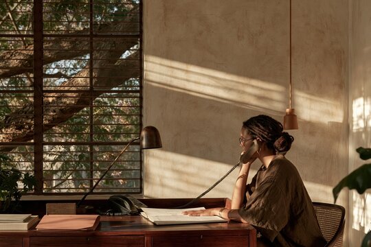 Black woman with locs talking on a desk phone in a sunlit office, natural mid-pitch moment with a blank press kit for creative public relations and career development portraits