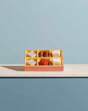 Assorted Indian sweets in an open gift box, featuring silver leaf kaju katli and saffron laddoo on a white stone counter with copy space for advertising