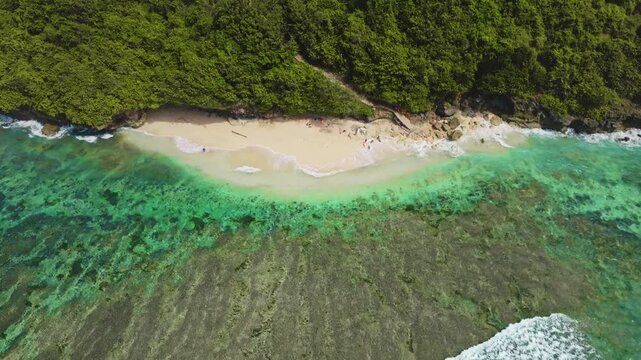 Bukit Peninsula coastline showing a sandy beach. Clear turquoise ocean water. Coral reef and vibrant green jungle cliffs creating a stunning tropical island landscape from an aerial drone perspective