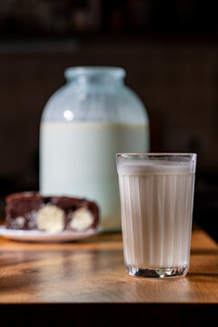 Fresh organic milk in faceted glass on wooden table. Glass of fresh whole milk closeup with rustic glass jar and delicious chocolate cake slice blurred in background on dark cozy kitchen counter