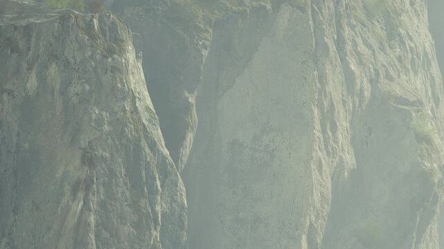 Thick fog covers tall cliffs by the ocean during early morning. The light creates a soft glow around the rocks while waves crash below. The view shows natural formations and mist.