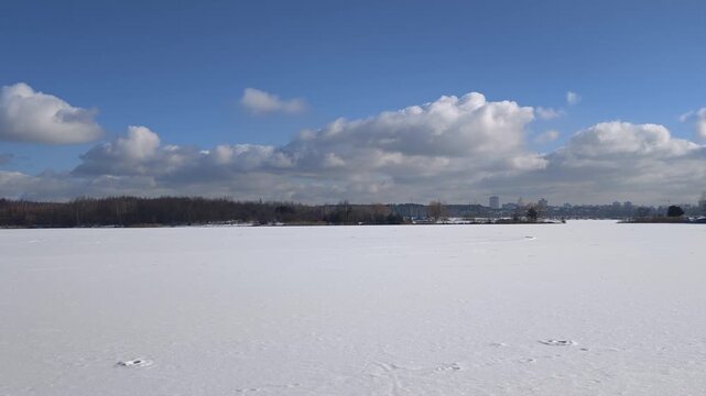 The ice on the lake and the grassy shores are covered with snow with footprints. On the far shore there are city ​​buildings and a mixed forest grows. Sunny winter weather and blue sky with clouds