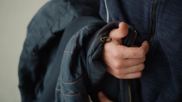 Close up of person holding sleeve and zipper area of two dark blue jackets focusing on hand grip and fabric texture. subject wears one jacket while grasping another with visible stitching