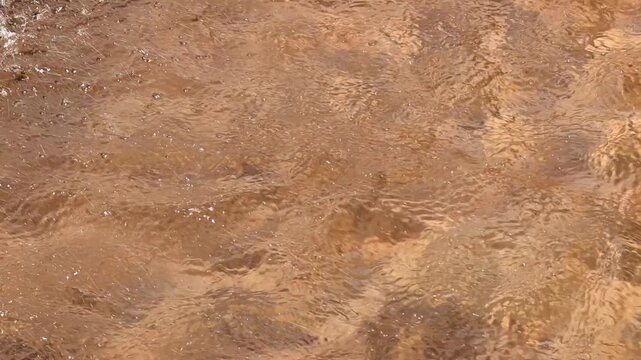Close-up view of water with brownish hues, showing delicate ripples and light reflections. The natural texture and movement of the liquid create an abstract pattern