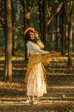 A young woman strolls calmly and casually through a field; she is wearing an open yellow shawl and a brown hat, with her arms spread wide. Boho style.
