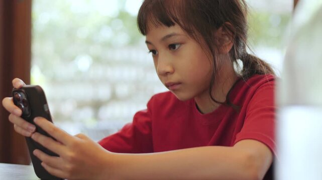 Close up Asian kid seriously focusing on a smartphone at a restaurant. Thai girl intently playing mobile games or watching content. Shot with selective focus and copy space. Digital lifestyle concept.