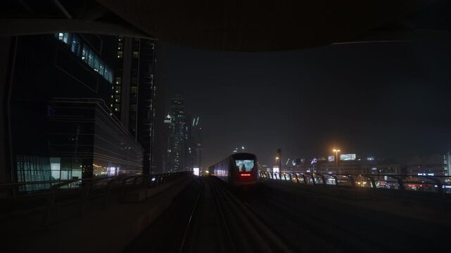 Passenger train recedes along an adjacent elevated railway as a front train POV travels toward illuminated city buildings at night
