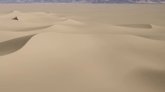 Waves of sand rise and fall in the desert under bright sunlight. The landscape shows vast, empty spaces with no signs of life or plant growth.