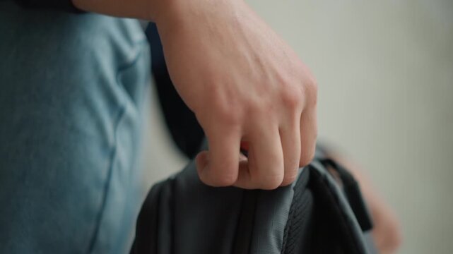 Close up of young man sitting and opening his gray backpack to retrieve items. Man is wearing blue jeans and dark sweatshirt with soft lighting highlighting texture of fabric materials