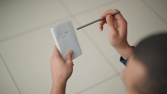 Close up of man holding open sketchbook with pencil drawing while examining it. notebook features spiral binding and blank pages set against soft indoor lighting on light colored floor tiles