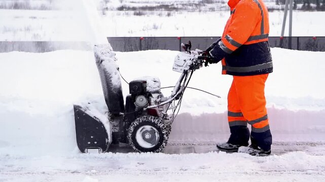 Worker clears snow with heavy-duty snow blower. Bright orange suit signals safety and visibility. Snow sprays forcefully into air behind machine. Scene shows winter cleanup effort in progress