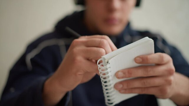 Close up of person holding and turning pages of spiral notebook with white paper focusing on hands interacting with book. Notebook is held in both hands while background remains softly