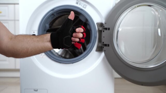 Hand in glove removes red garment from washing machine. Person shows clean laundry inside machine. Modern appliance in bright kitchen setting. Perfect for laundry, home cleaning