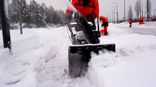 Snowplow operator clears icy street in winter. Workers in bright orange gear assist nearby. Snow falls steadily around cleared roadway. Scene shows active winter maintenance effort
