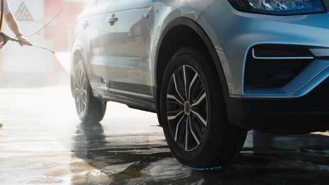 Person sprays water onto silver car's side panel. Water jets splash, glistening wet ground. Vehicle's sleek design reflects spray mist. Cleaner's motion highlights routine care