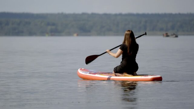 Woman kneels on red paddleboard, paddling calm lake. She grips paddle, focused on smooth water. Distant trees line horizon under soft sky. Serene paddleboarding moment, quiet and calm